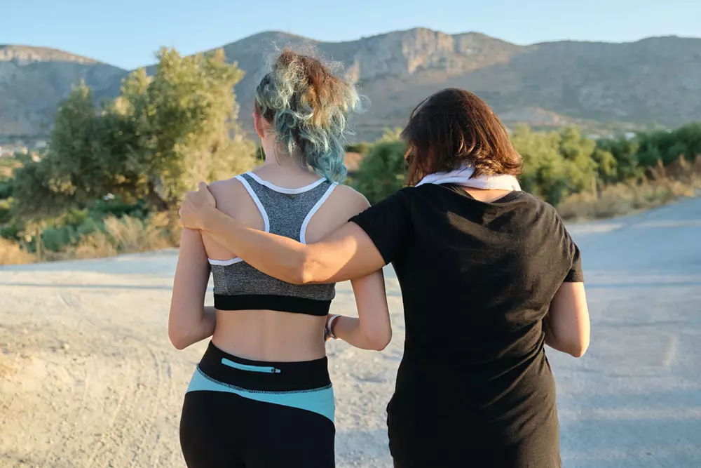 Joven y su entrenadora de espaldas. La entrenadora le pasa el brazo por el hombre y la arropa.